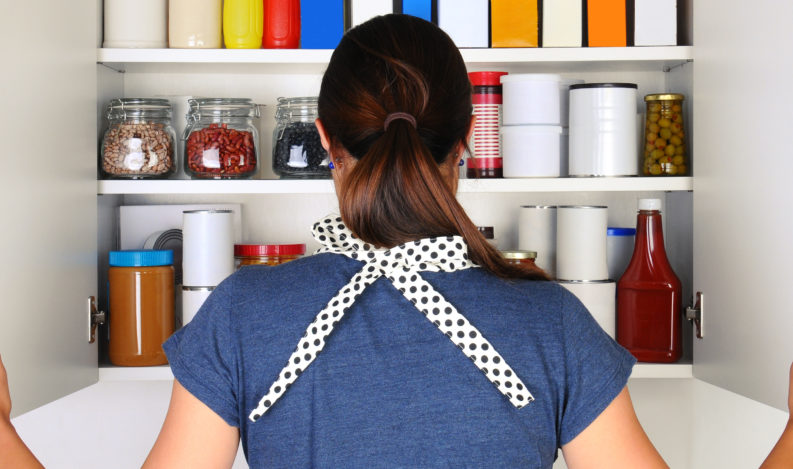 A woman seen from behind opening the doors to a fully stocked pantry. The cupboard is filled with various food stuff and groceries all with blank labels. Horizontal format, the woman is unrecognizable.
