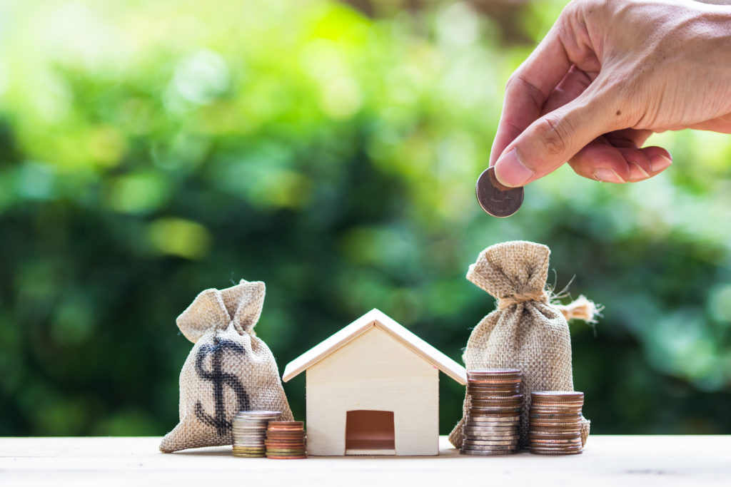 Saving money, home loan, mortgage, a property investment for future concept : A man hand putting money coin over small residence house and money bag with nature background. A sustainable investment.