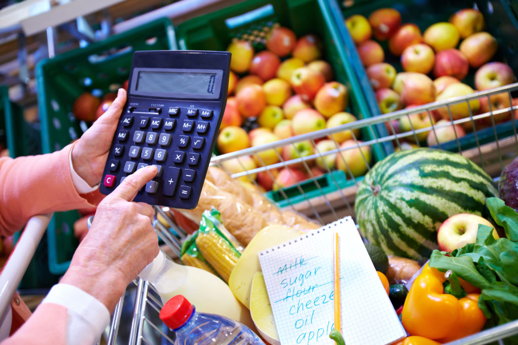 Image of senior woman hand touching buttons of calculator with goods in cart near by