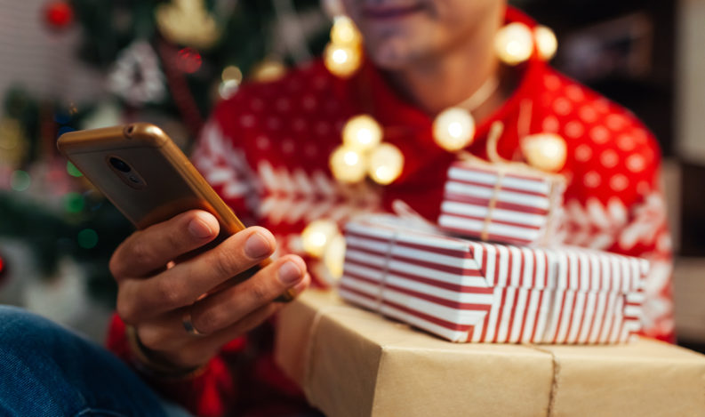 Christmas shopping online. Man buying New year presents using smartphone. Guy holding gift boxes and phone by Christmas tree