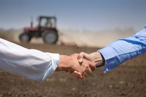 Two businessmen shaking hands in field with tractor working in background. Agribusiness concept