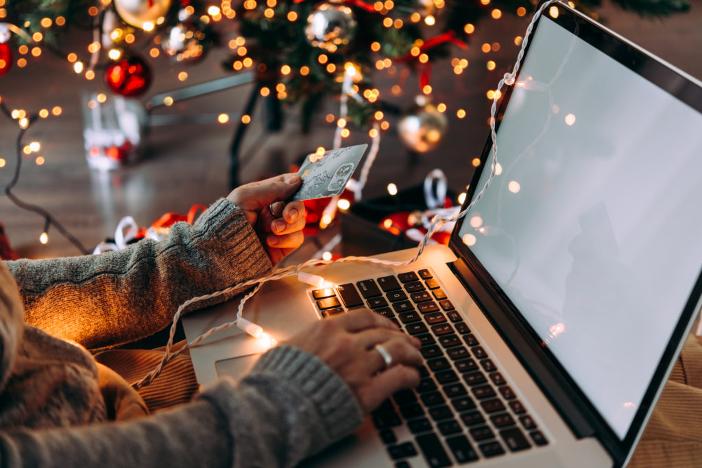 Christmas shopping online. Women's hands typing on laptop while holding up a credit card. Christmas tree and lights in the background.