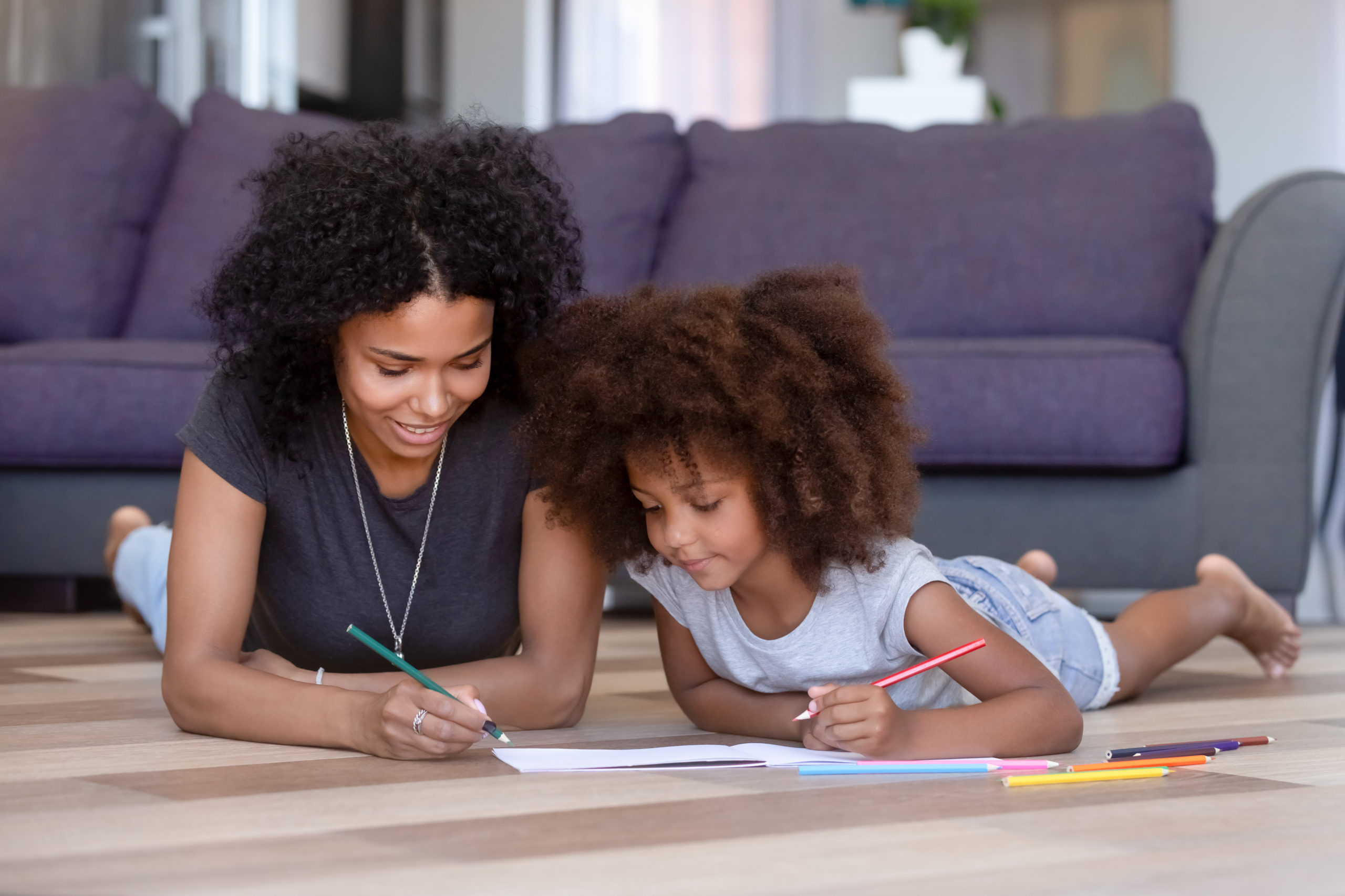 African American mother coloring with daughter, lying on warm floor in living room at home, smiling family spending free time, weekend together, woman drawing colored pencils with pretty girl
